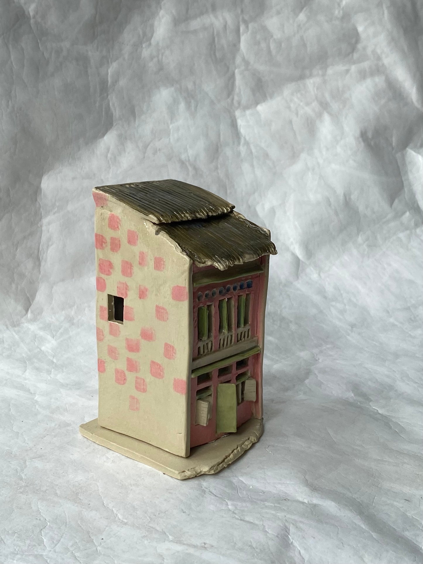 A small handcrafted ceramic of Singapore shophouse with a beige body, pink checkered pattern, and a dark brown roof, set against a white paper background.