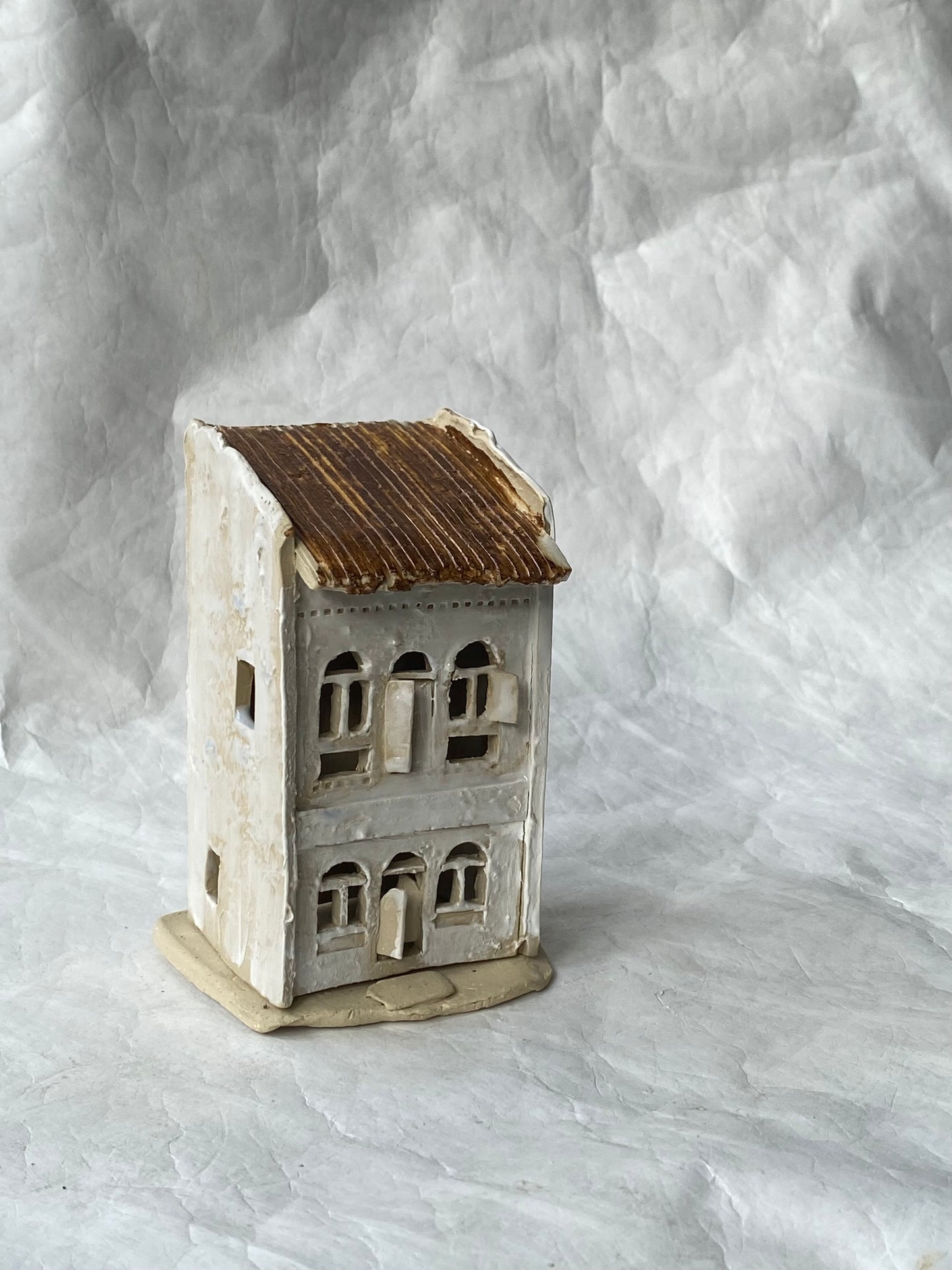 A small handmade ceramic house figurine with a weathered off-white body, a rustic brown roof, and a beige base, set against a wrinkled white paper backdrop.