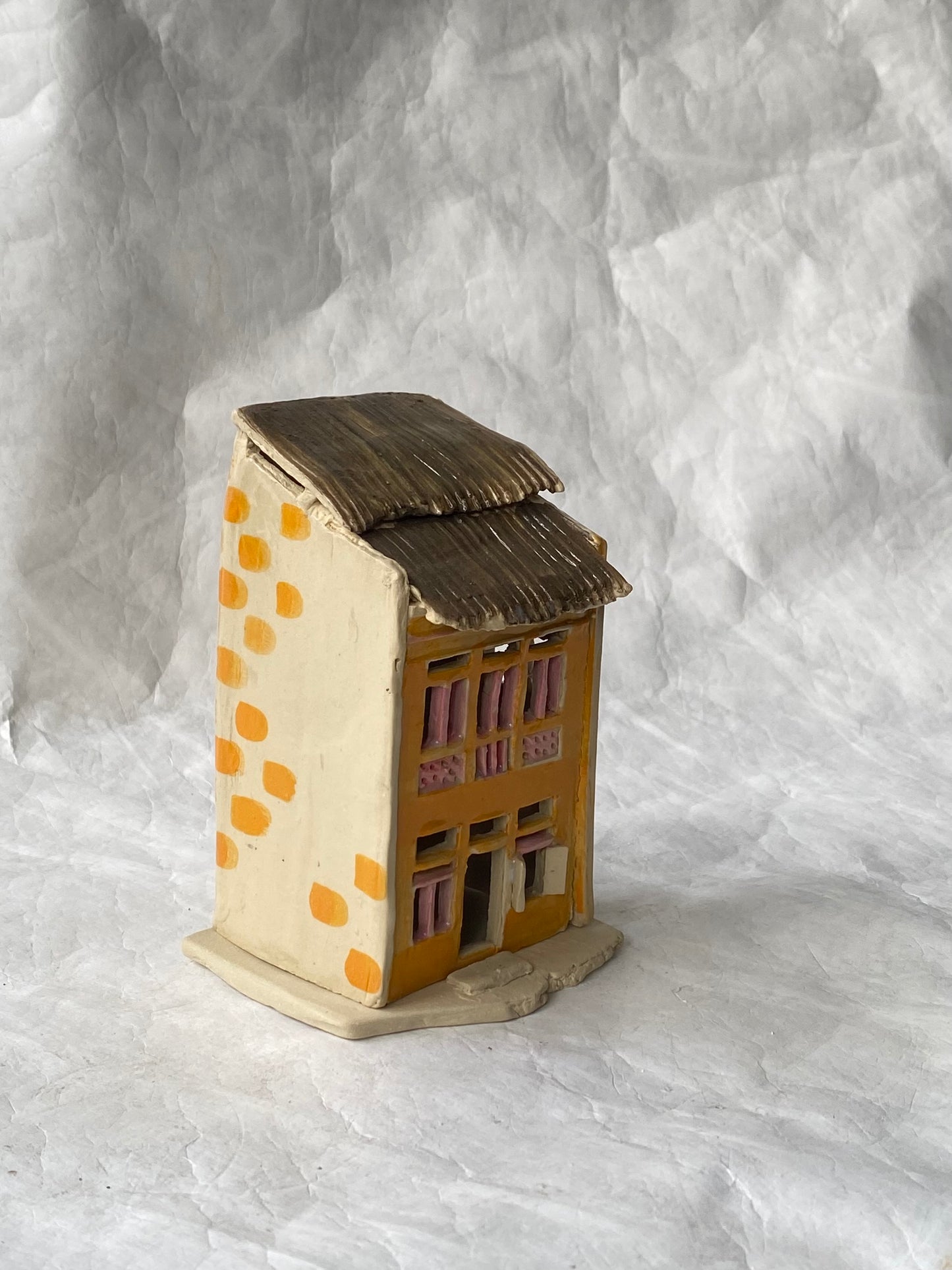 A small hand-painted ceramic house figurine with a beige-coloured body, orange dots, a brown roof, and pink window details, set against a wrinkled white paper background.
