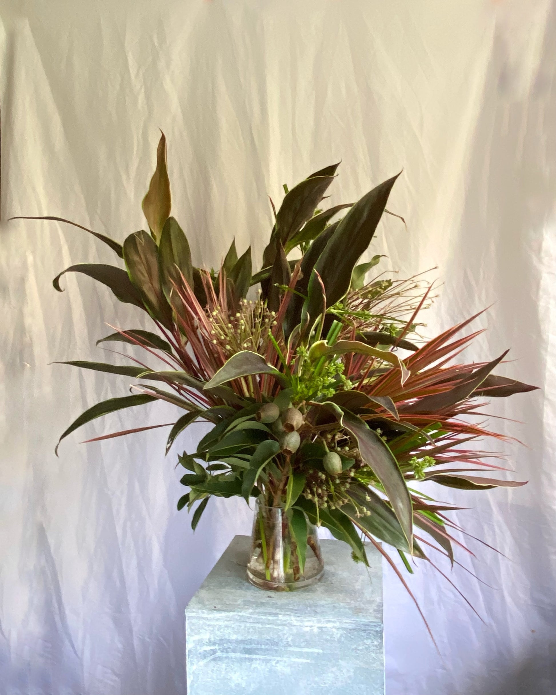 A sculptural botanical arrangement by foRM of Mondays, showcasing an elegant mix of handpicked green leaves and foliage in a clear glass vessel against a minimalist background.