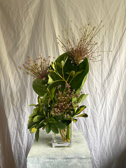 A sculptural botanical arrangement by foRM of Mondays, showcasing an elegant mix of handpicked green leaves and foliage in a clear glass vessel against a minimalist background.