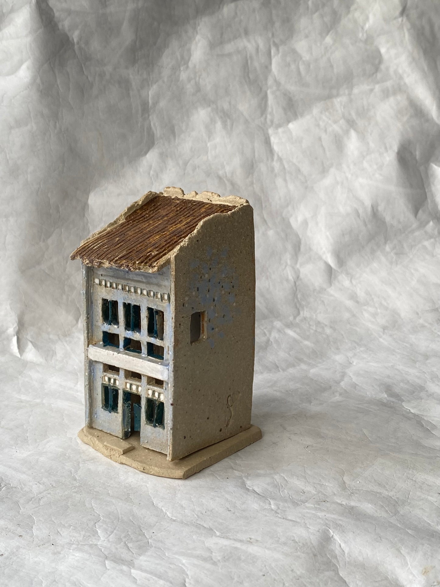 A small handbuilt ceramic of Singapore shophouse figurine with a weathered blue grey body, dark brown roof, and blue window accents, set against a  white paper background.