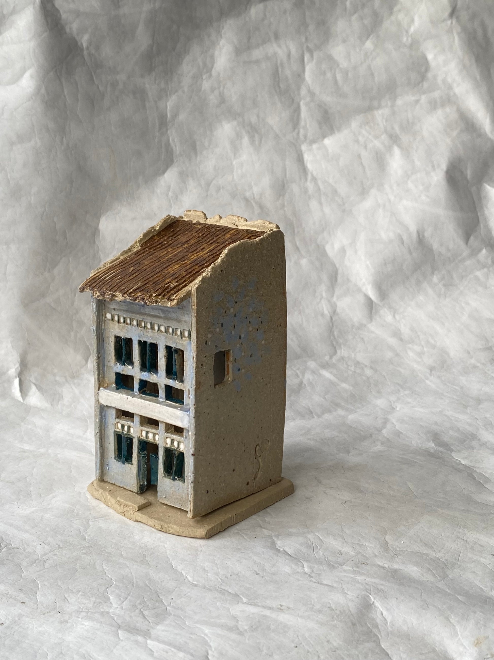 A small handbuilt ceramic of Singapore shophouse figurine with a weathered blue grey body, dark brown roof, and blue window accents, set against a  white paper background.