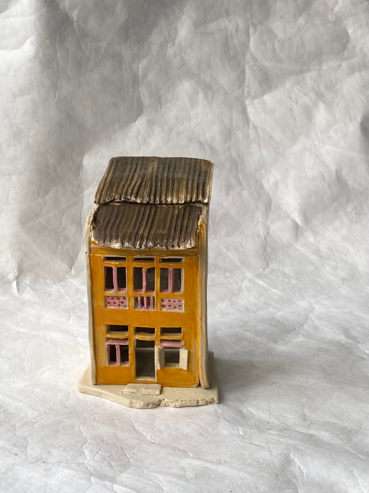 A small handbuilt ceramic of Singapore shophouse figurine with a yellow-orange facade, brown roof, and pink window accents, standing on a textured white background.