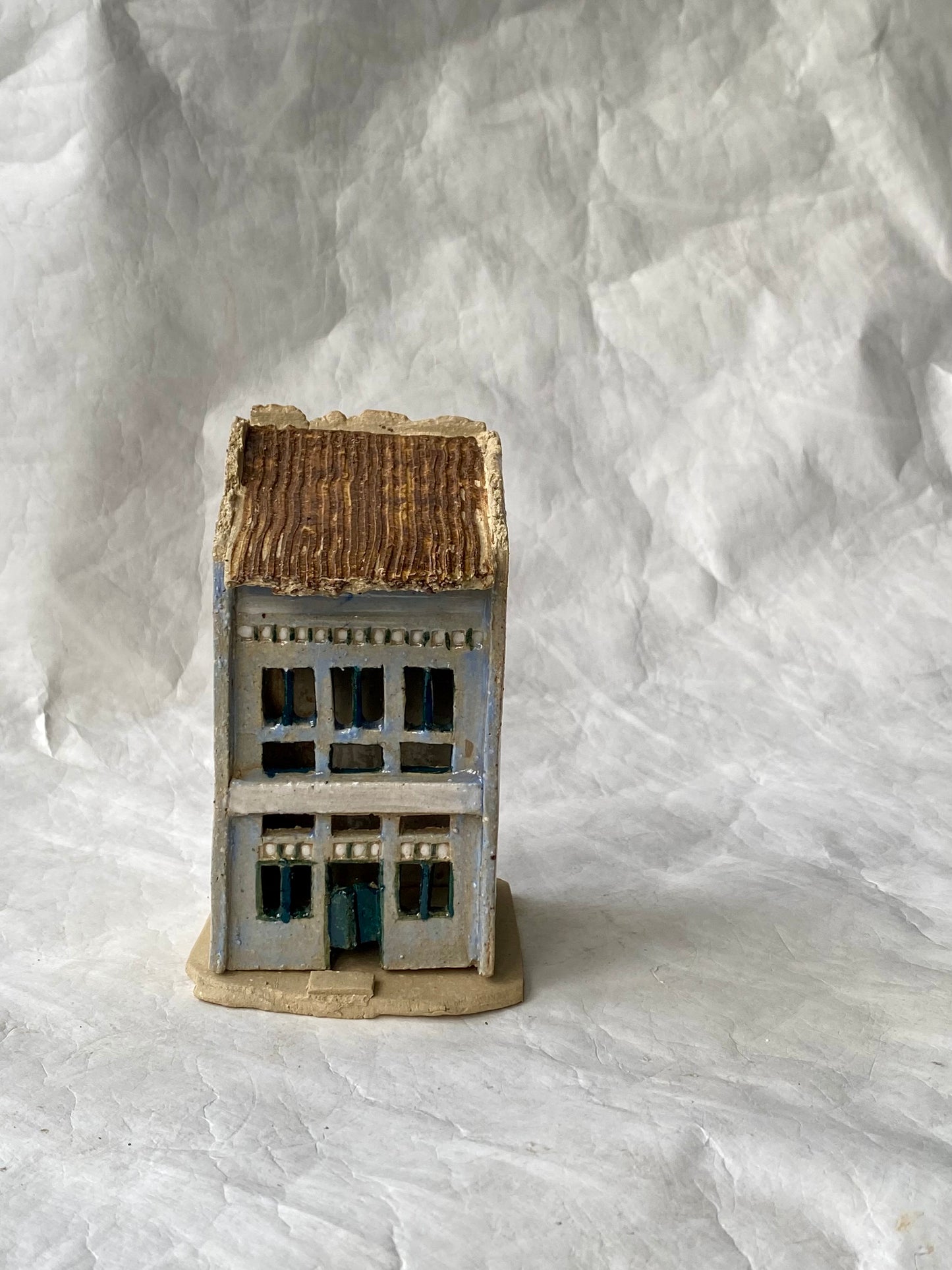 A miniature model of a two-story building with a weathered brown roof, blue-gray walls, and dark blue window details, standing on a textured beige base against a white backdrop.