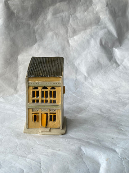 A handbuilt ceramic of Singapore shophouse with a dark gray ribbed roof and beige/yellow facade, placed on a white paper background.