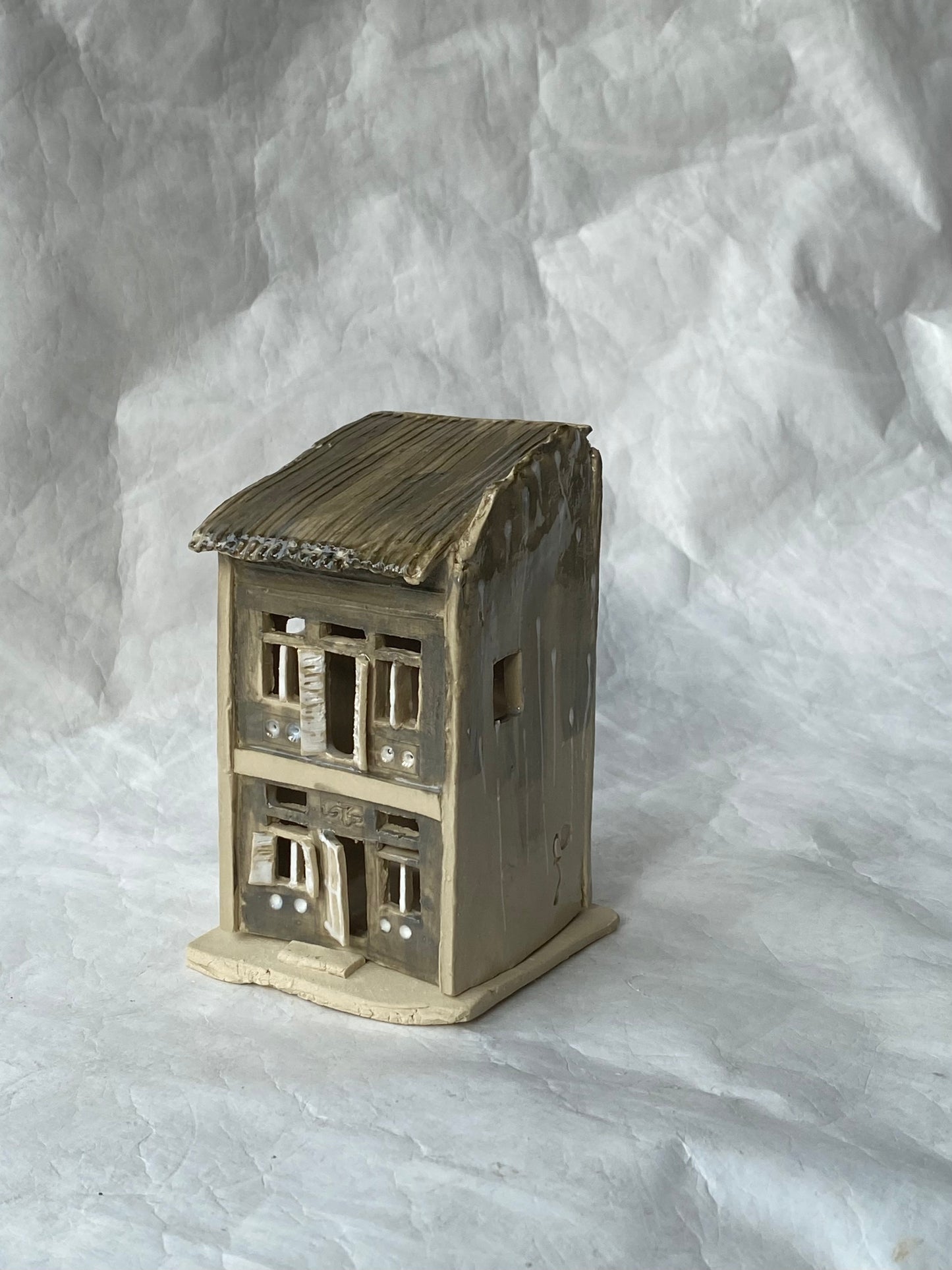A handbuilt ceramic of Singapore shophouse with a dark ribbed roof and light grey facade, placed on a white paper background.
