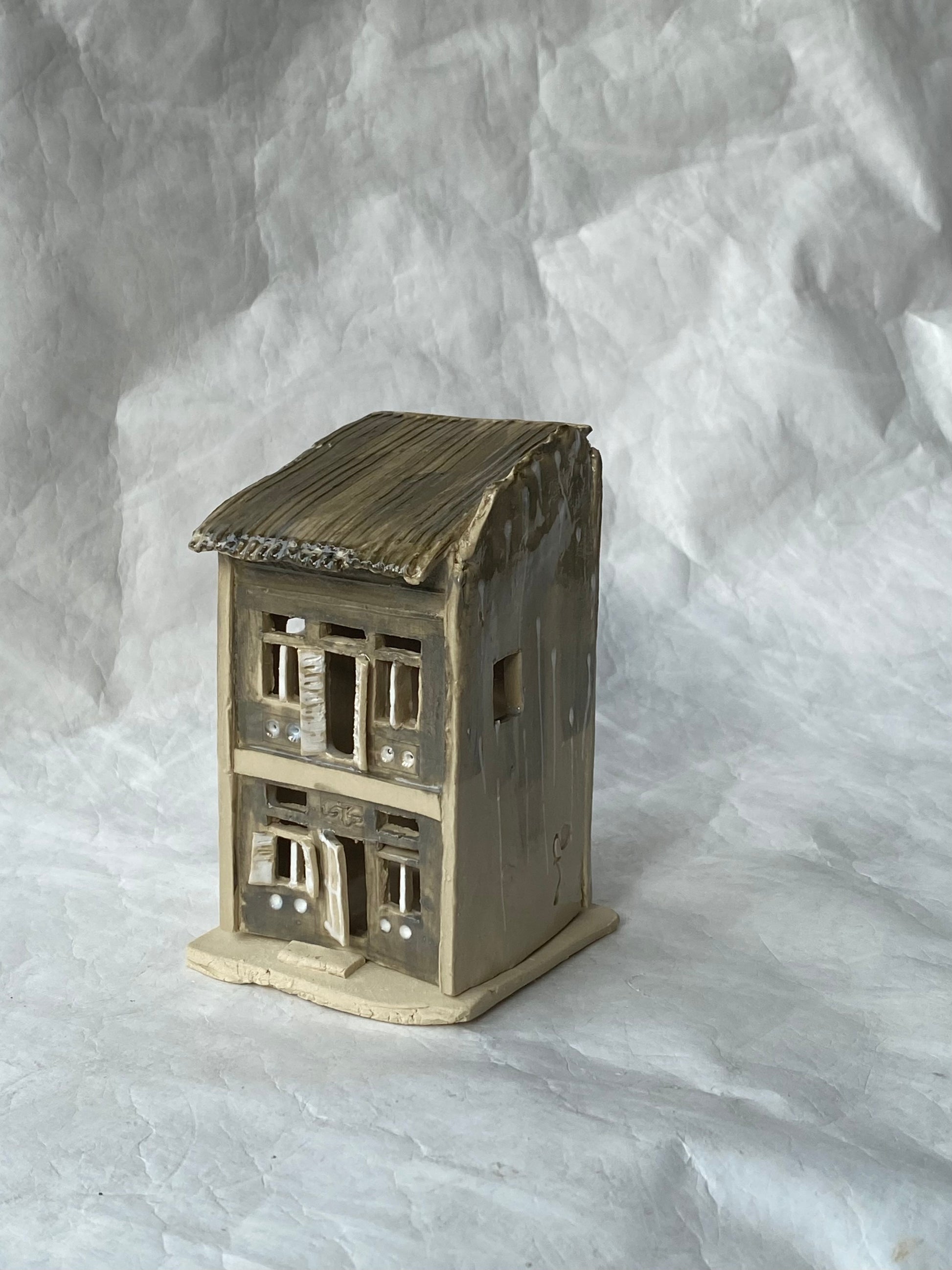 A handbuilt ceramic of Singapore shophouse with a dark ribbed roof and light grey facade, placed on a white paper background.
