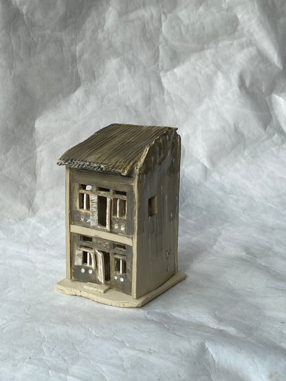 A handbuilt ceramic of Singapore shophouse with a dark ribbed roof and light grey facade, placed on a white paper background.
