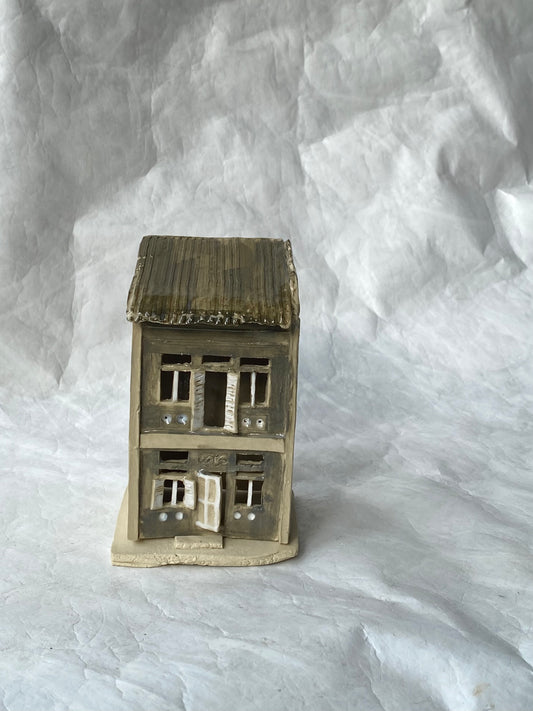 A handbuilt ceramic of Singapore shophouse with a dark ribbed roof and light grey facade, placed on a white paper background.
