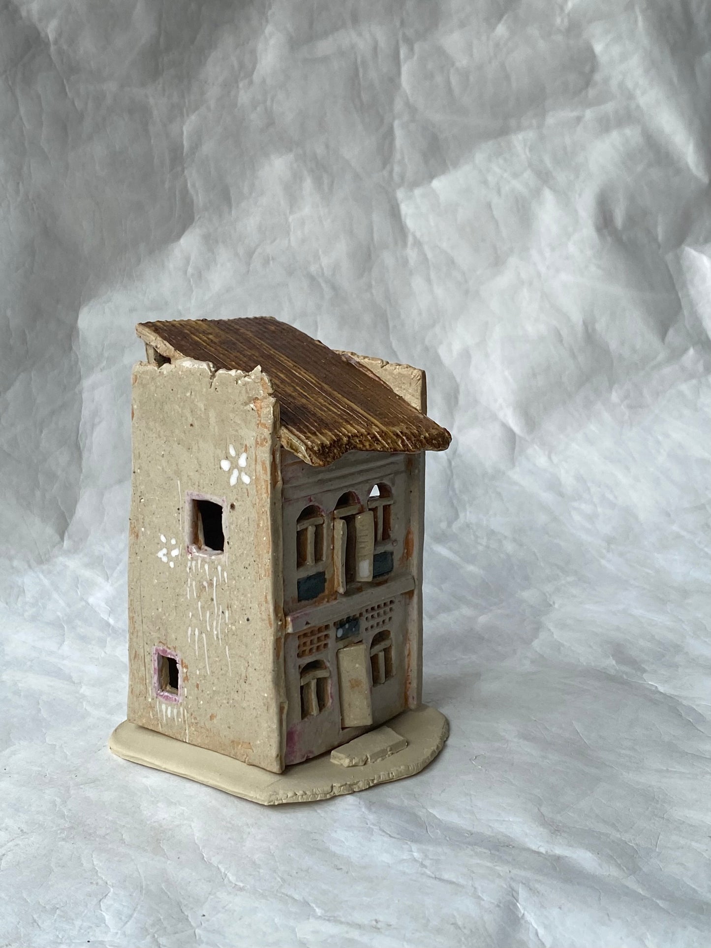 A small handmade ceramic of Singapore shophouse figurine with a weathered pinkish grey body, brown roof, and detailed window patterns, set against a  white paper backdrop.