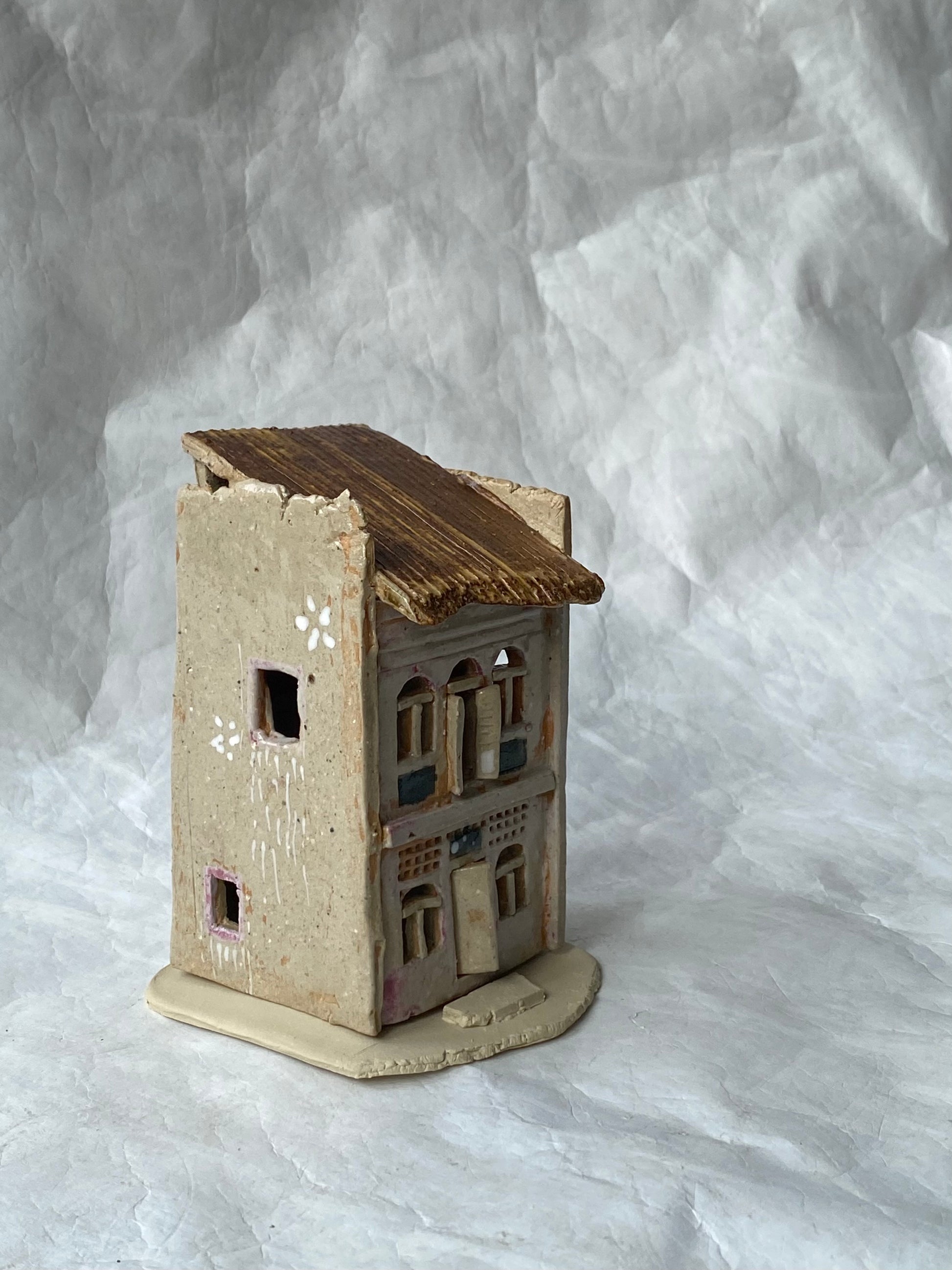 A small handmade ceramic of Singapore shophouse figurine with a weathered pinkish grey body, brown roof, and detailed window patterns, set against a  white paper backdrop.