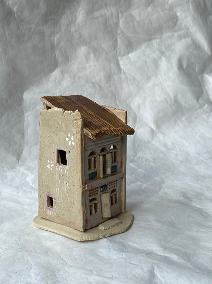 A small handmade ceramic of Singapore shophouse figurine with a weathered pinkish grey body, brown roof, and detailed window patterns, set against a  white paper backdrop.