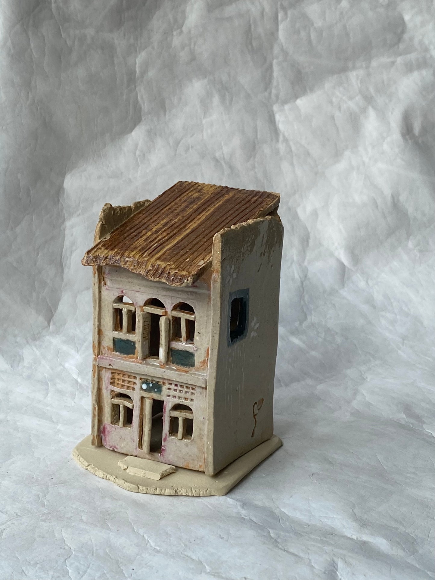 A miniature handcrafted clay building of Singapore shophouse with a weathered brown roof, pinkish grey walls, and painted window details, set against a wrinkled white paper backdrop.
