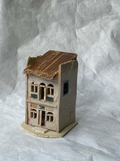 A miniature handcrafted clay building of Singapore shophouse with a weathered brown roof, pinkish grey walls, and painted window details, set against a wrinkled white paper backdrop.