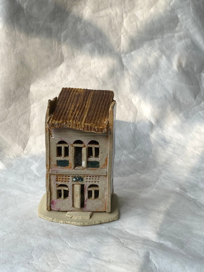 A hand built ceramic of Singapore shophouse figurine with a brown roof and a pinkish grey facade, displayed on a textured white background.