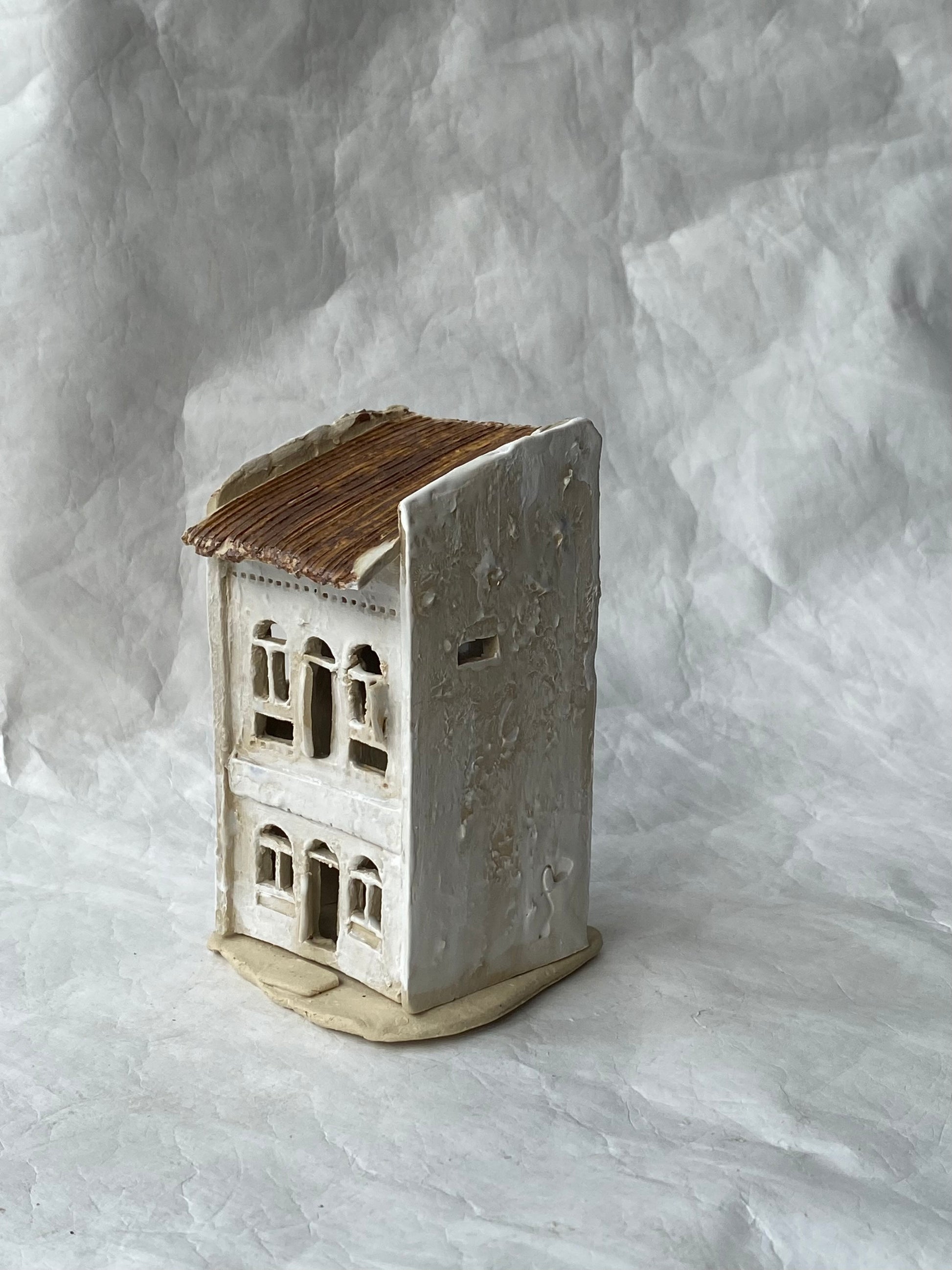 A small handmade ceramic of Singapore shophouse figurine with a textured off-white body, brown roof, and beige base, photographed against a  light gray backdrop.