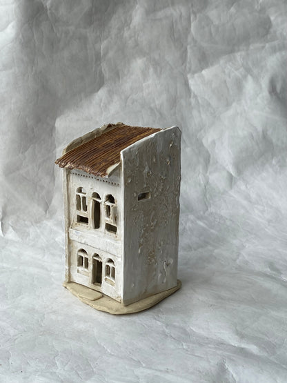 A small handmade ceramic of Singapore shophouse figurine with a textured off-white body, brown roof, and beige base, photographed against a  light gray backdrop.