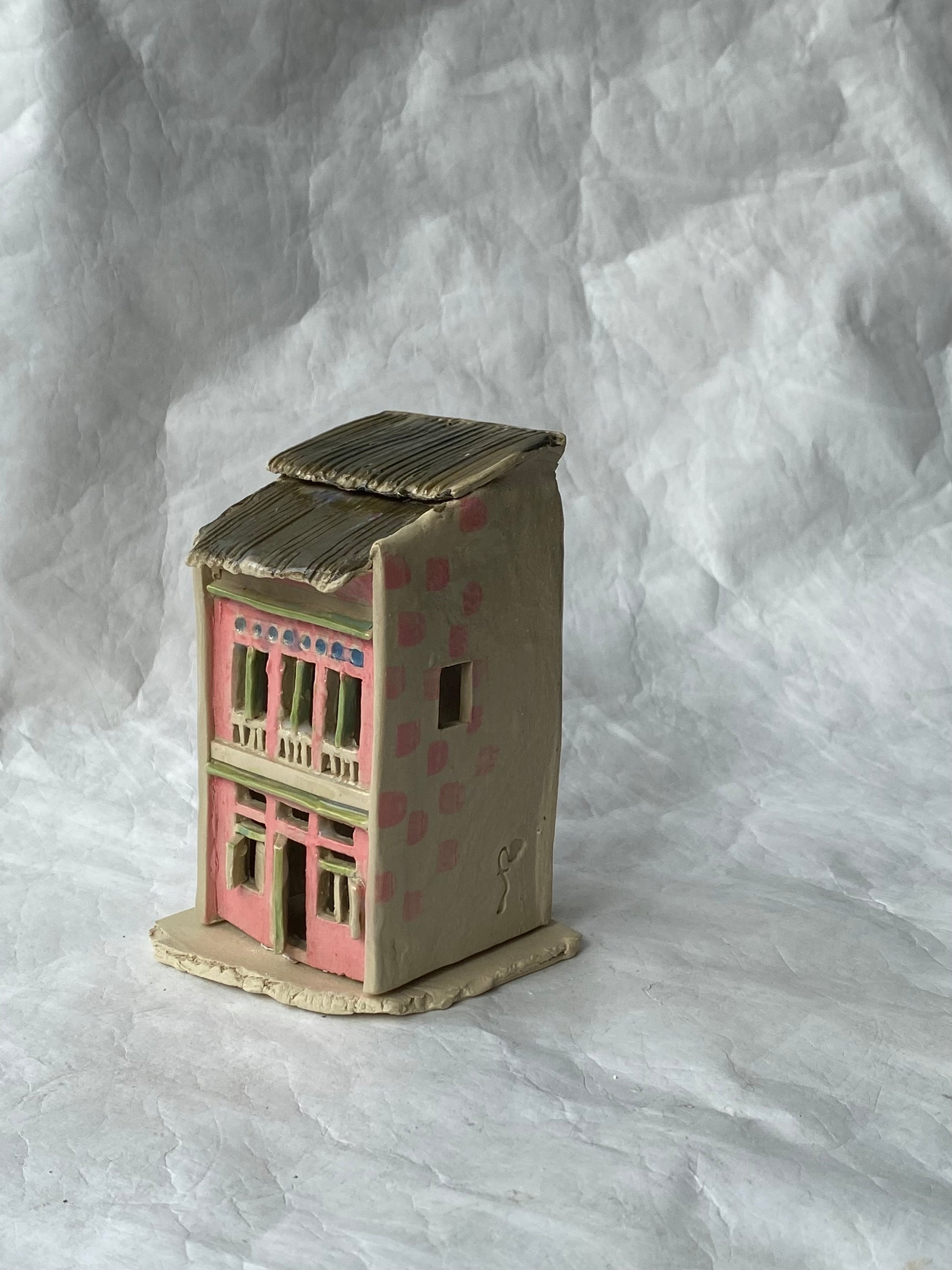 A small handcrafted clay house figurine with a pink facade, beige walls, and a brown roof, set against a wrinkled white paper backdrop.