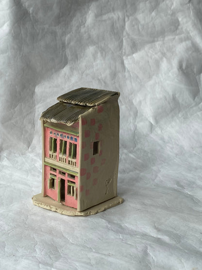 A small handcrafted clay house figurine with a pink facade, beige walls, and a brown roof, set against a wrinkled white paper backdrop.