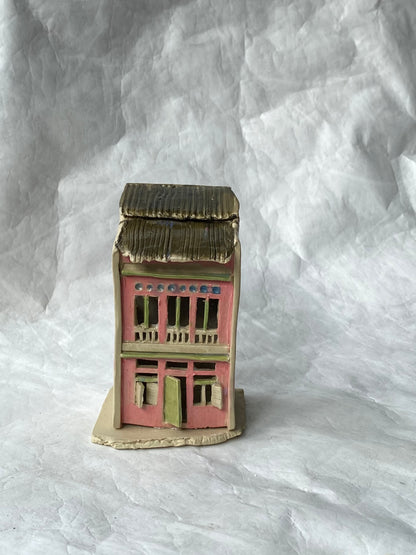 A miniature handcrafted clay of Singapore shophouse figurine with a pink facade, green trim, and a dark brown roof, set against a white background.