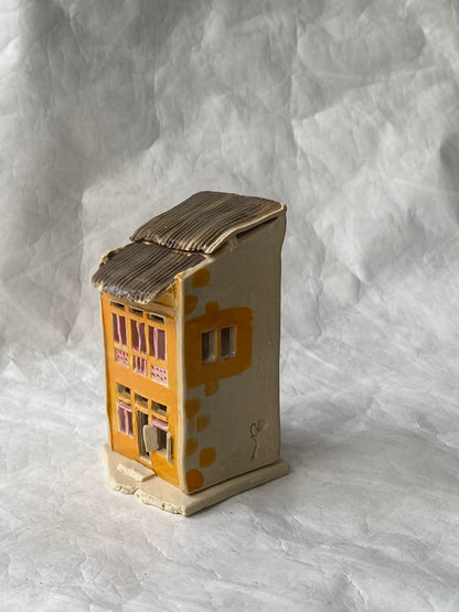 A small hand-painted ceramic Singapore shophouse figurine with a brown textured roof, yellow-orange facade, and beige base, set against a white backdrop.