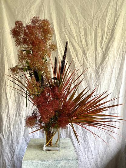 A sculptural botanical arrangement titled Just Another Lovely Day by foRM of Mondays, showcasing an elegant mix of handpicked interesting foliage and grass  in a clear glass vessel against a minimalist background.
