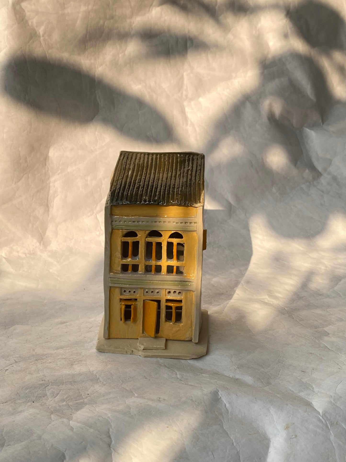 A handbuilt ceramic of Singapore shophouse with a dark gray ribbed roof and beige/yellow facade, placed on a white paper background.