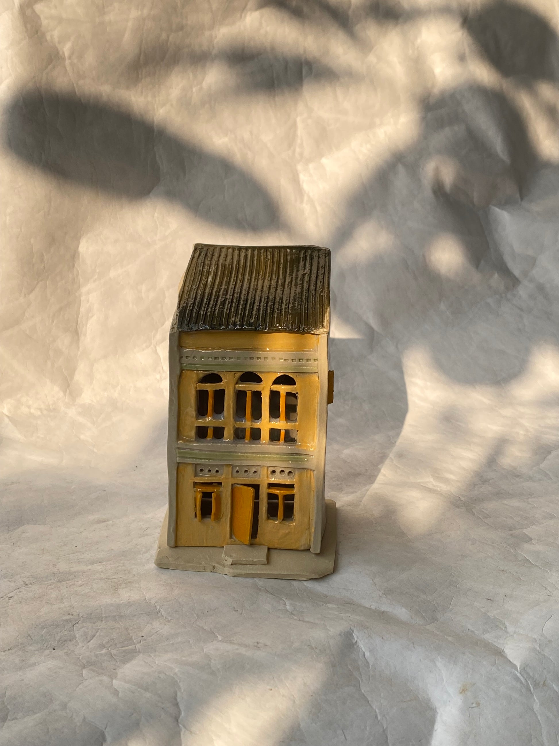 A handbuilt ceramic of Singapore shophouse with a dark gray ribbed roof and beige/yellow facade, placed on a white paper background.