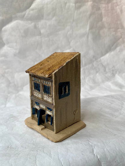 A small handcrafted clay miniature of a Singapore heritage shophouse with a brown roof, beige walls, and blue window accents, displayed on a textured white background.