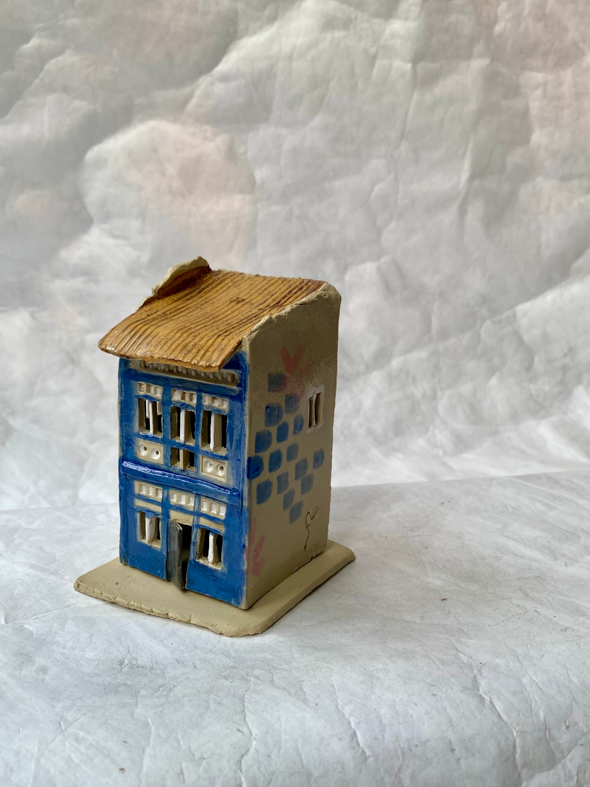 A small handcrafted ceramic shophouse figurine by foRM of Mondays with a blue front facade, beige side walls, and a brown textured roof, displayed on a  white fbackground.