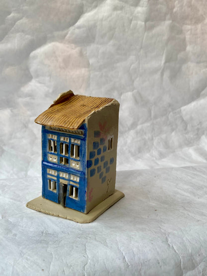 A small handcrafted ceramic shophouse figurine by foRM of Mondays with a blue front facade, beige side walls, and a brown textured roof, displayed on a  white fbackground.