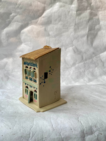 A small handcrafted clay house figurine of Singapore shophouse with a beige body, brown roof, and light blue and green decorative details, set against a white background.