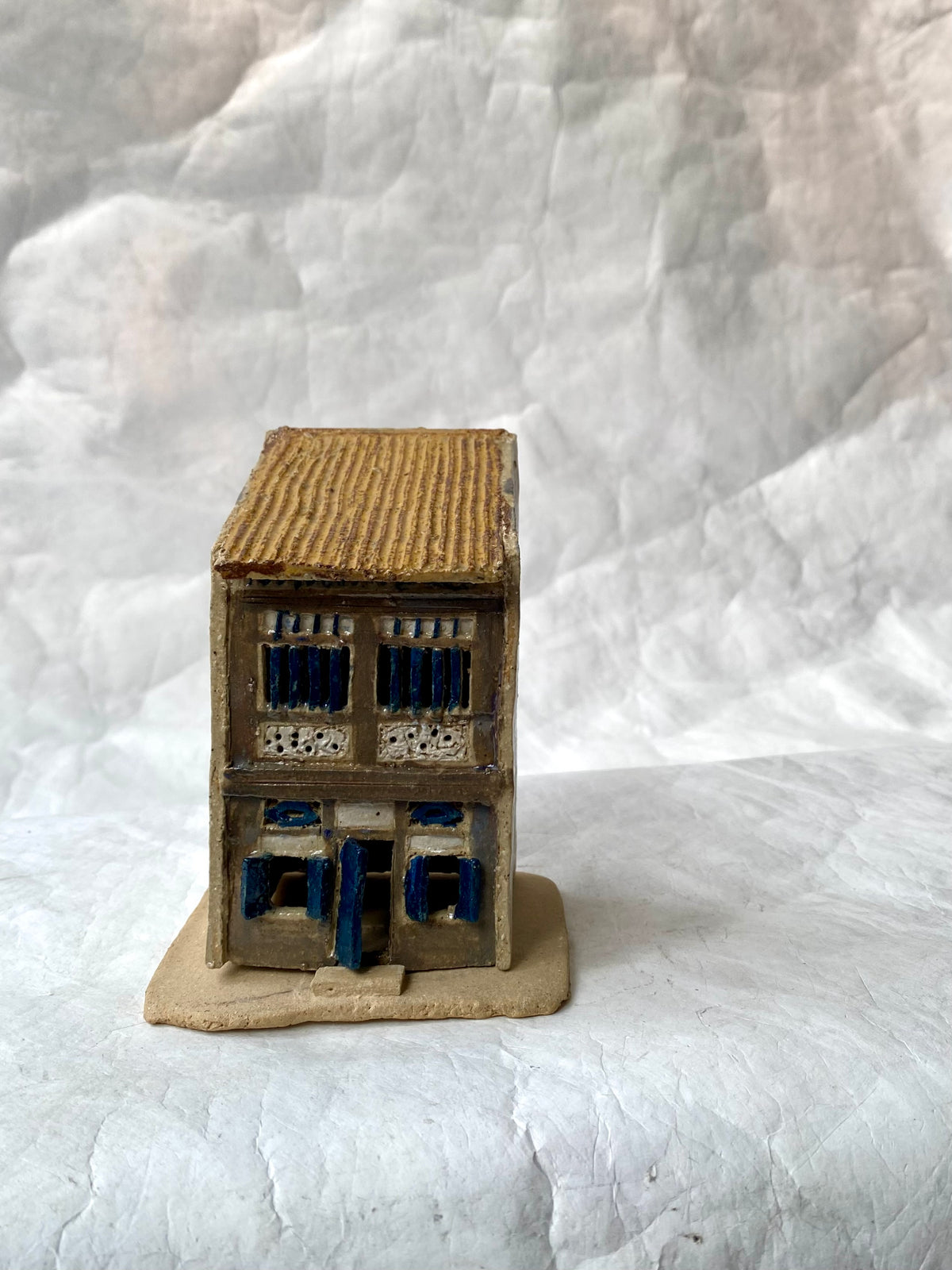 A handbuilt ceramic of Singapore shophouse with brown ribbed roof and dark gray/blue facade, placed on a white paper background.