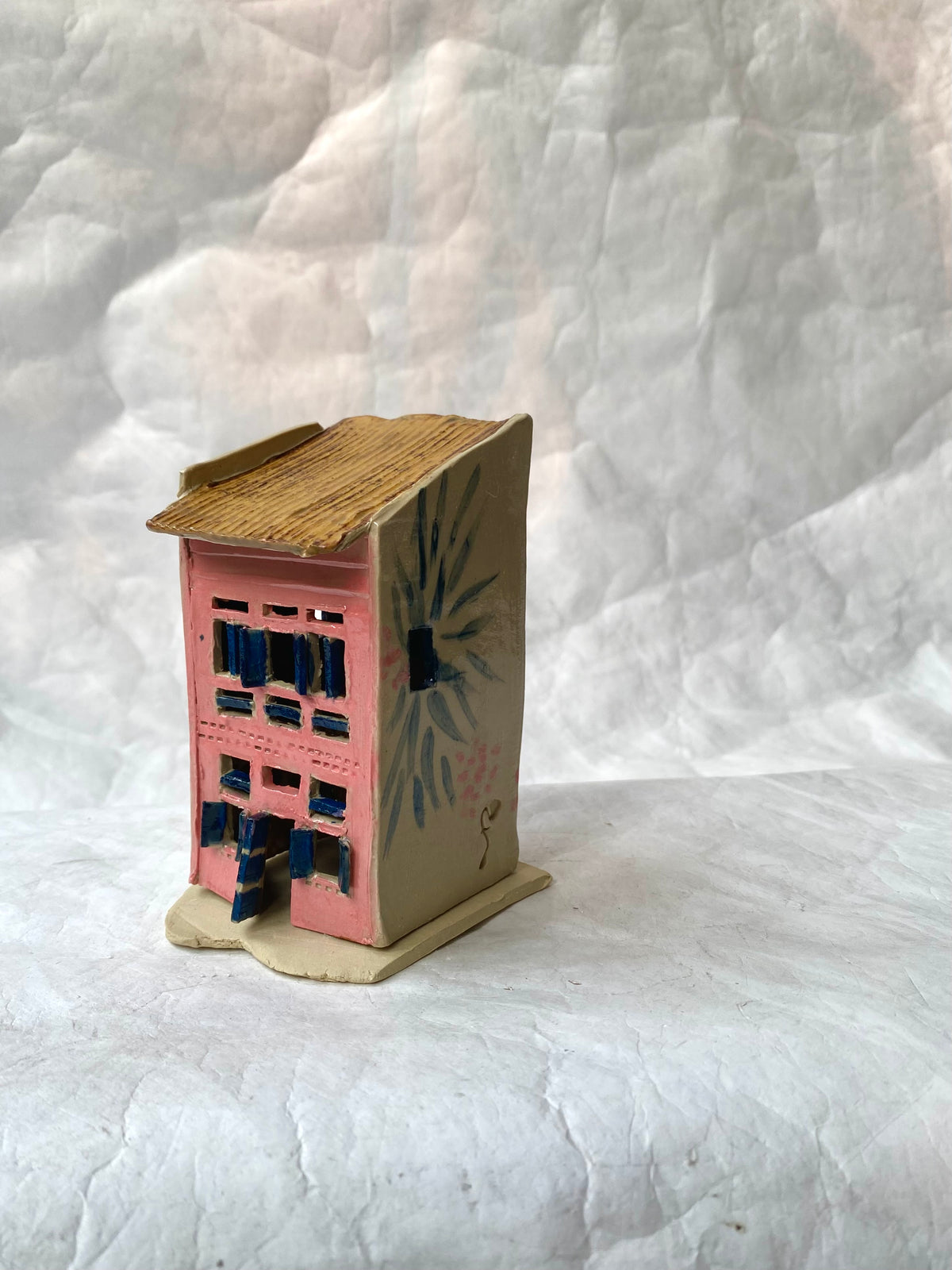 A small handcrafted Singapore shophouse figurine with a pink facade, blue window details, a brown roof, and a beige base, set against a wrinkled white fabric backdrop.