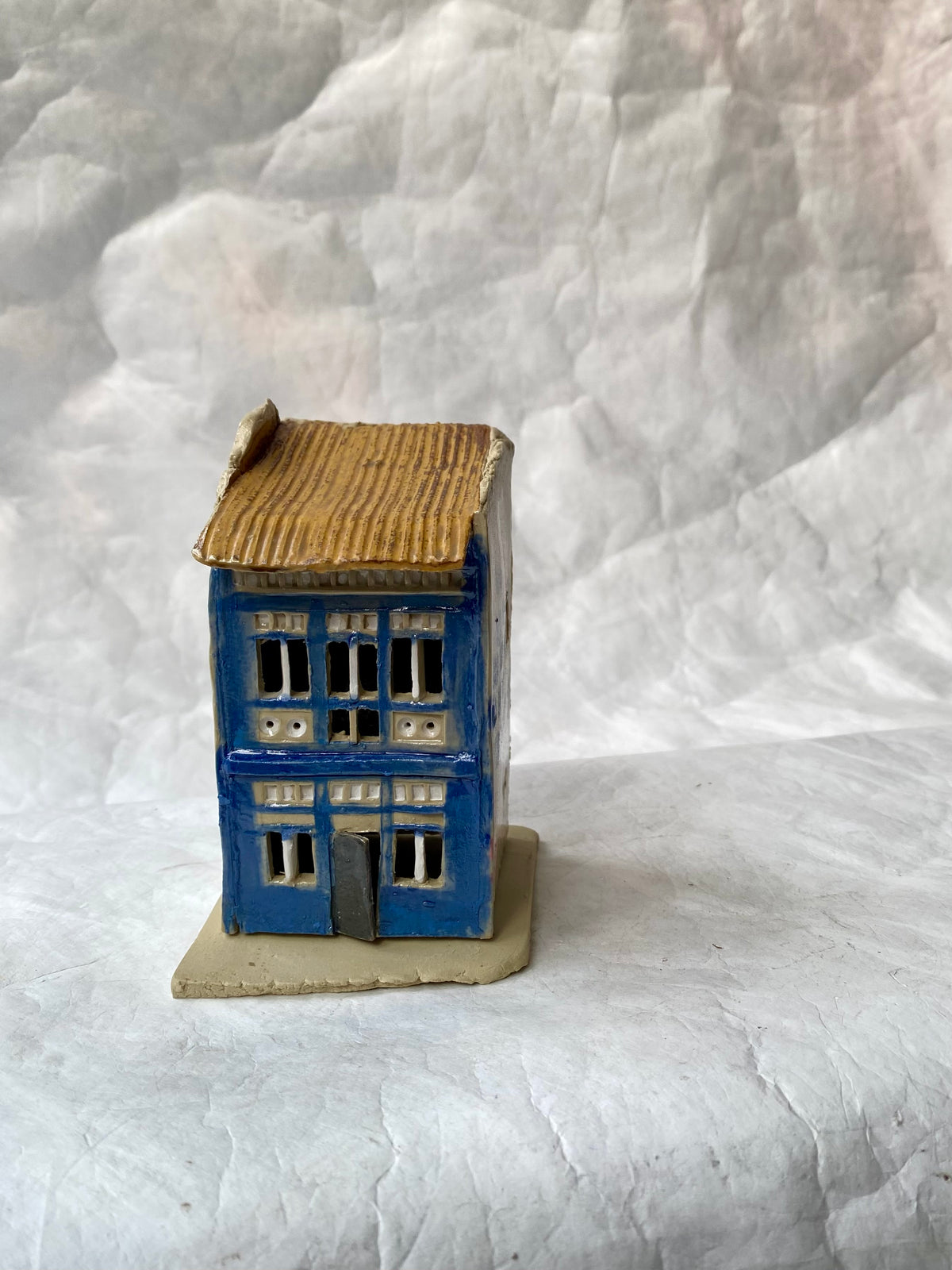 A handbuilt ceramic of Singapore shophouse with a brown ribbed roof and blue facade, placed on a white paper background.