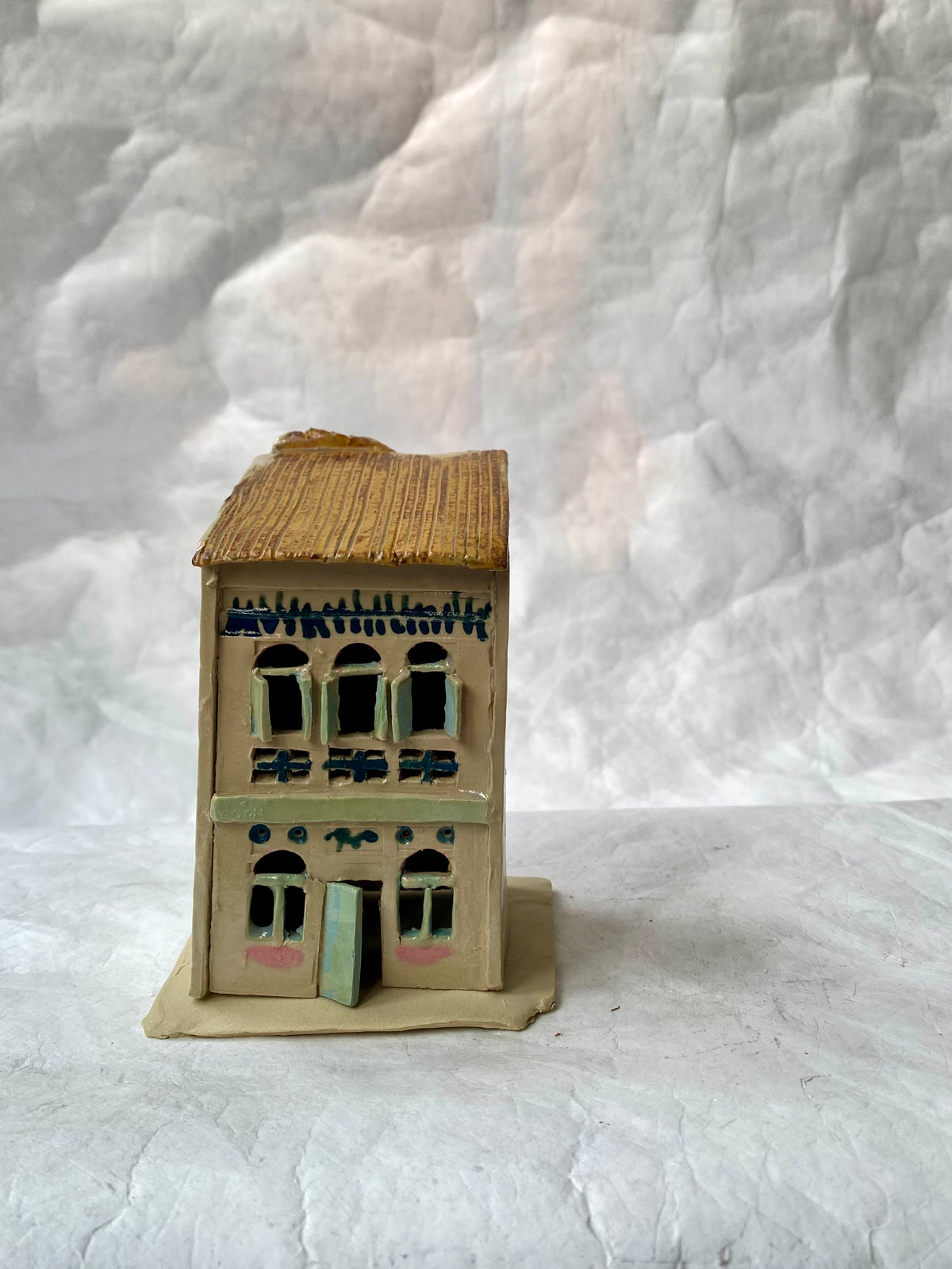 A handbuilt ceramic of Singapore shophouse with a brown ribbed roof and beige/pastels facade, green door, placed on a white paper background.