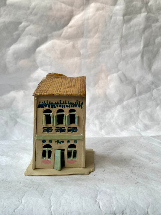 A handbuilt ceramic of Singapore shophouse with a brown ribbed roof and beige/pastels facade, green door, placed on a white paper background.