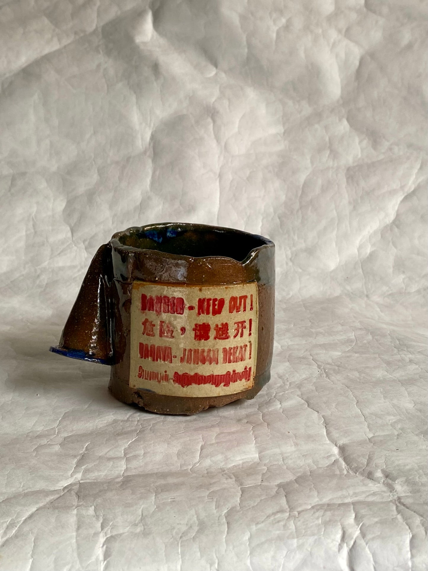 A handbuilt brown ceramic mug with a cone handle, featuring a red warning label that reads 'DANGER - KED OUT!' Set against grey background.