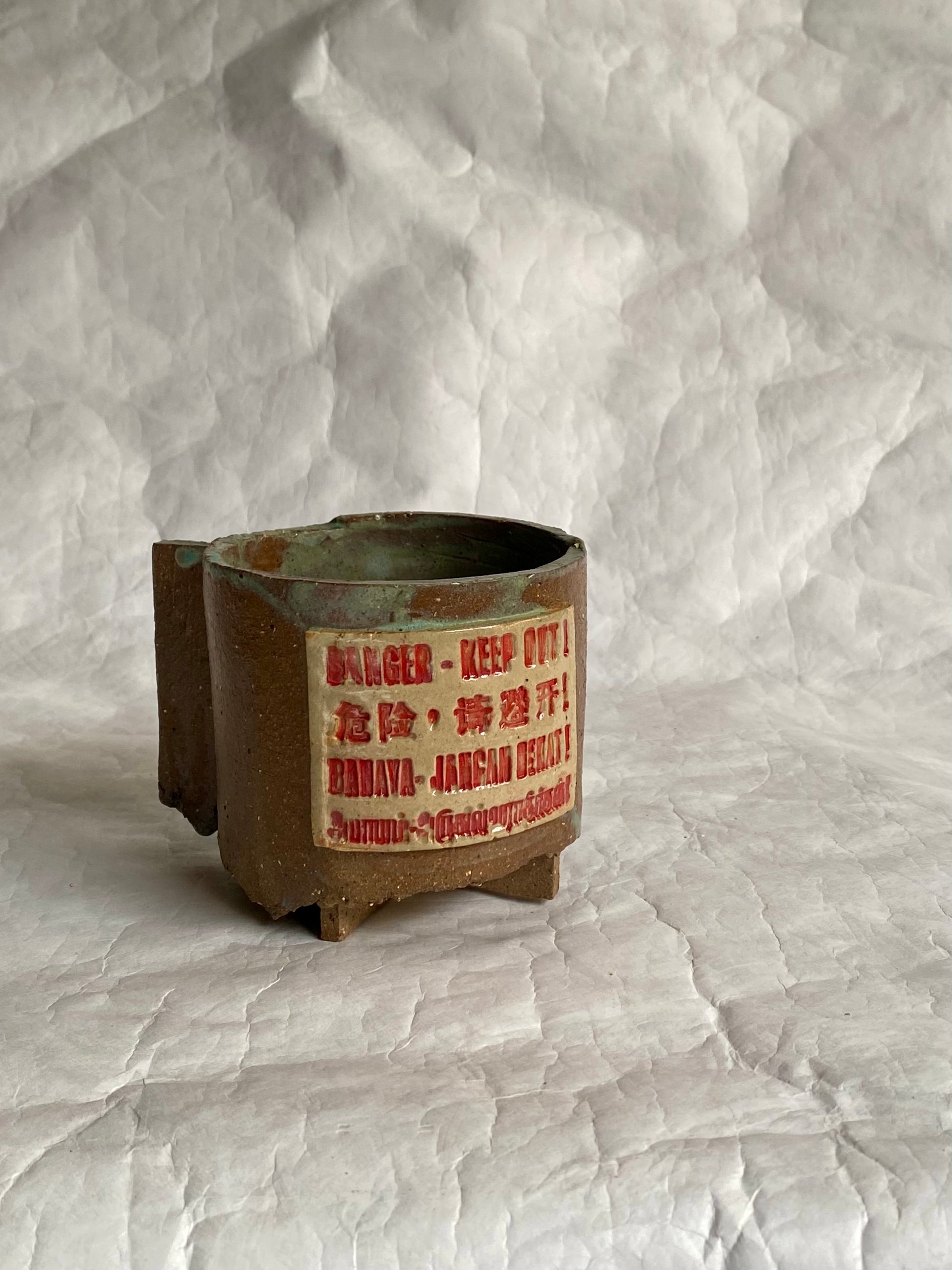 A handbuilt brown ceramic mug with a red warning label reading 'DANGER - KEEP OUT!' and additional text in Chinese and English, set against a  white paper background.