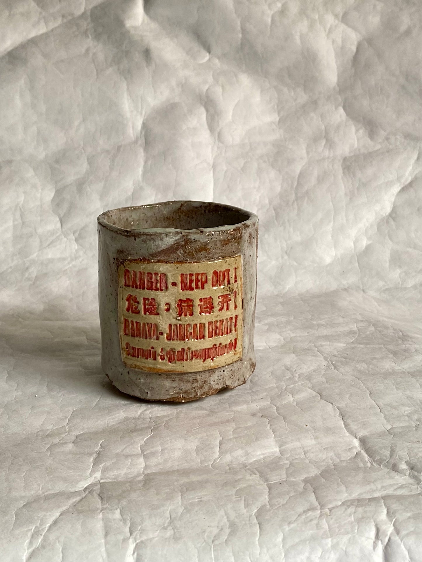 A handbuilt ceramic cup with a distressed, weathered finish, featuring a central label with red text “Danger- Keep out” signage and Chinese characters. The item is photographed on a  white paper  backdrop.