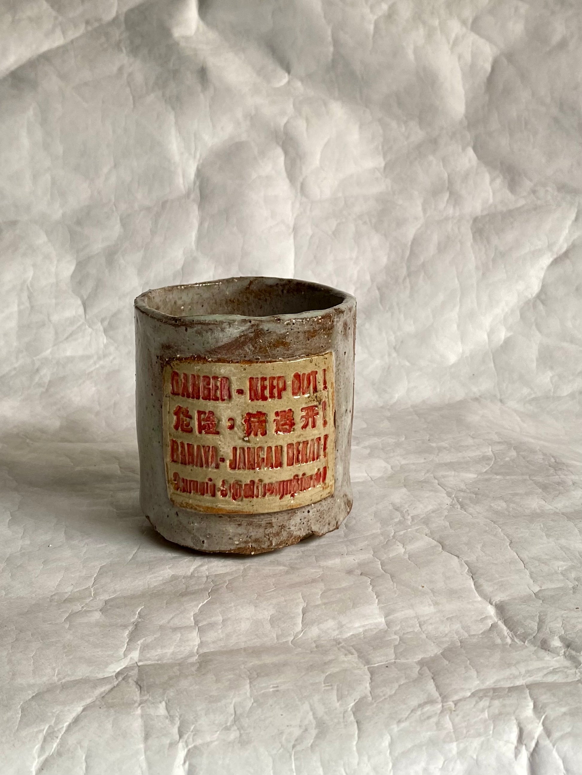 A handbuilt ceramic cup with a distressed, weathered finish, featuring a central label with red text “Danger- Keep out” signage and Chinese characters. The item is photographed on a  white paper  backdrop.