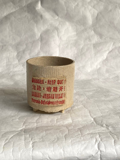 A handbuilt ceramic cup or tumbler in a light beige stoneware finish, featuring red printed warning text that reads 'DANGER - KEEP OUT!' along with additional text in Chinese and English. The cup is photographed on a white paper background.