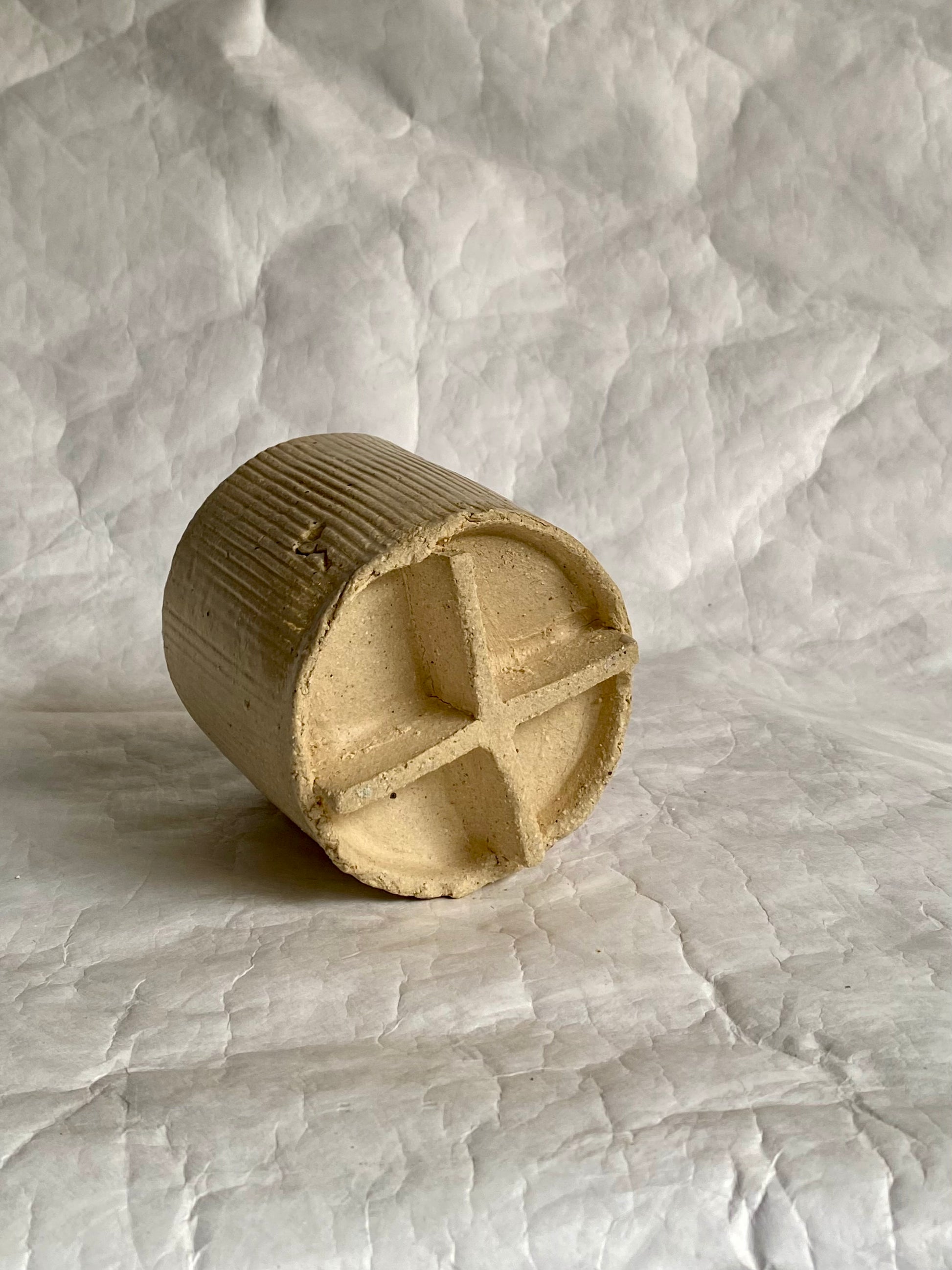 Details of a handbuilt ceramic cup with a ribbed cylindrical body and a cross-shaped bottom on a textured white paper  background