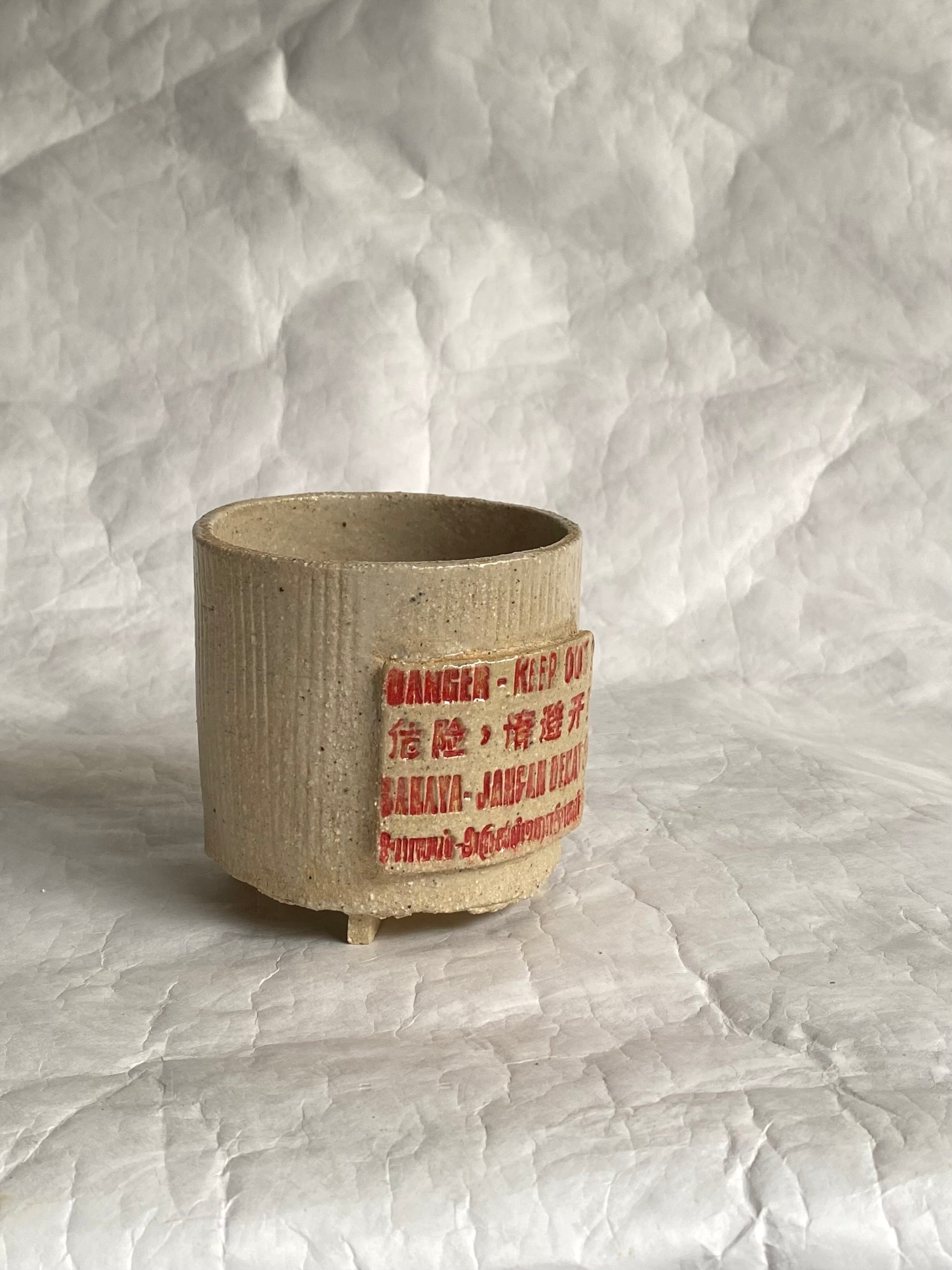A handmade ceramic cup with a textured beige body and a red printed  label, set against a white paper backdrop.