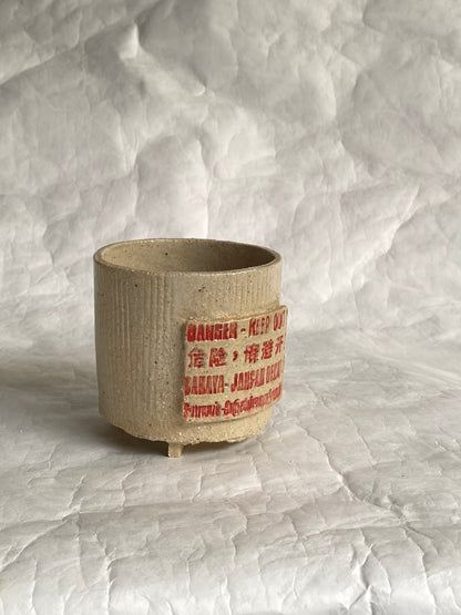 A handmade ceramic cup with a textured beige body and a red printed  label, set against a white paper backdrop.